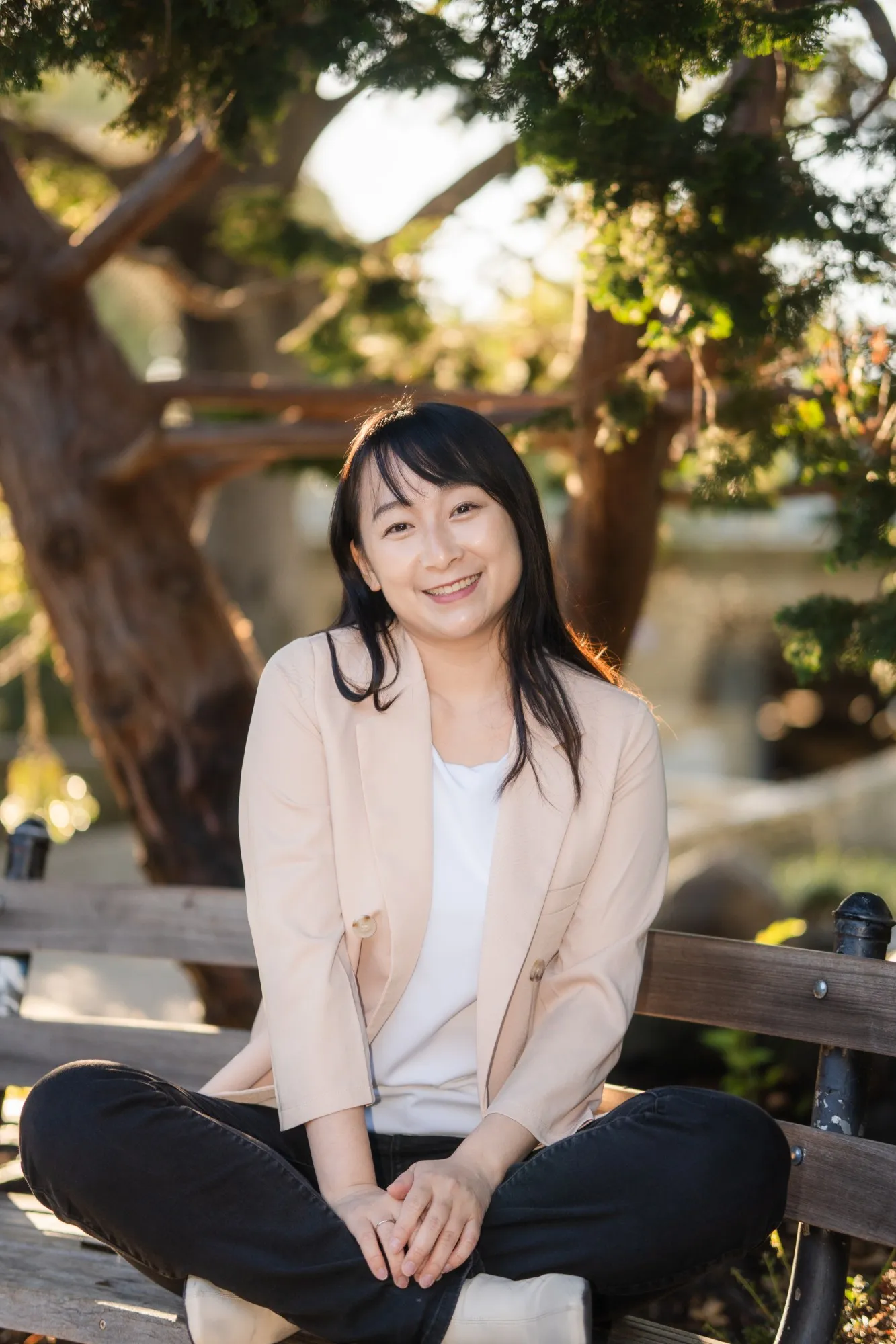 Bilingual therapist Zi Zheng smiling, sitting cross-legged on a bench under a tree